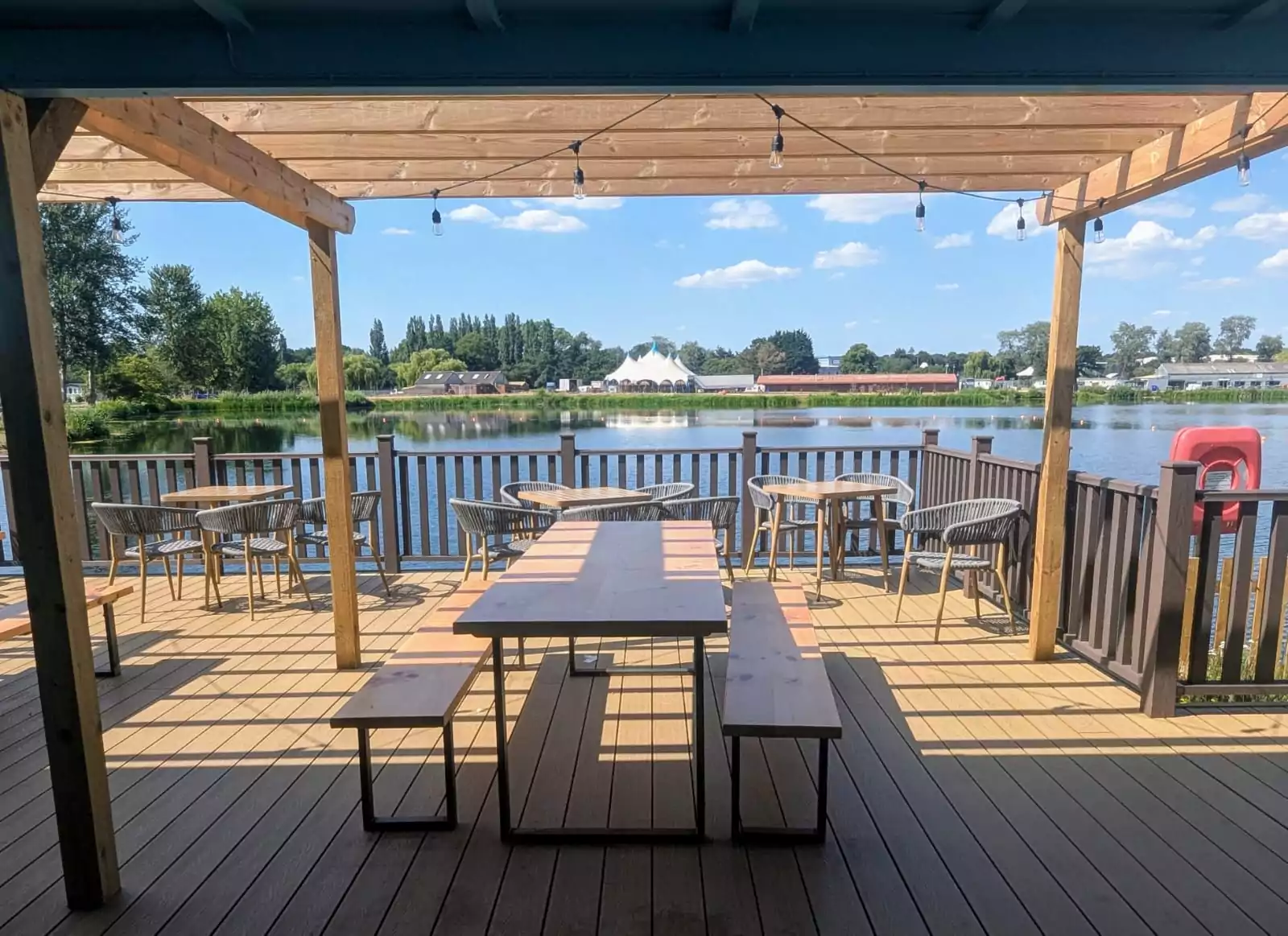Outdoor dining deck overlooking a calm lake with trees and buildings in the distance under a sunny sky.