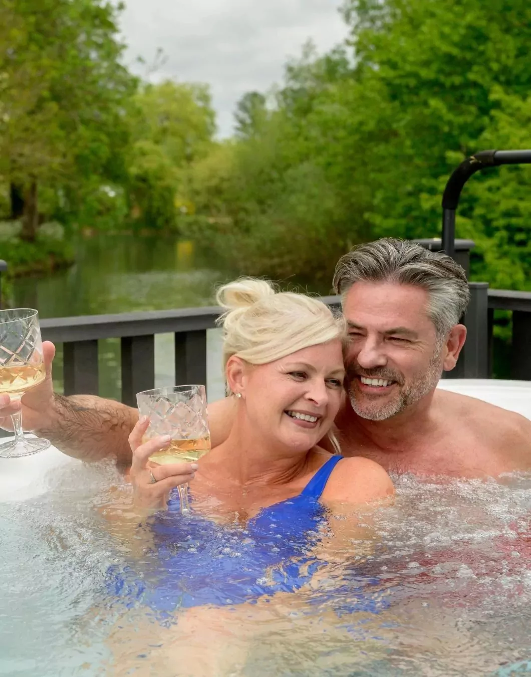Smiling couple relaxing in a hot tub at Billing Aquadrome, holding glasses of white wine with a scenic river and greenery in the background