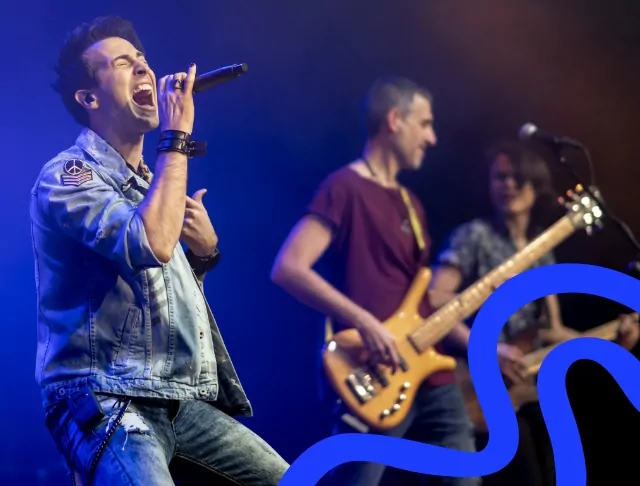 A passionate lead singer performing on stage, wearing a denim jacket and singing into a microphone. Behind him, a bass guitarist and an electric guitarist play under blue stage lighting.
