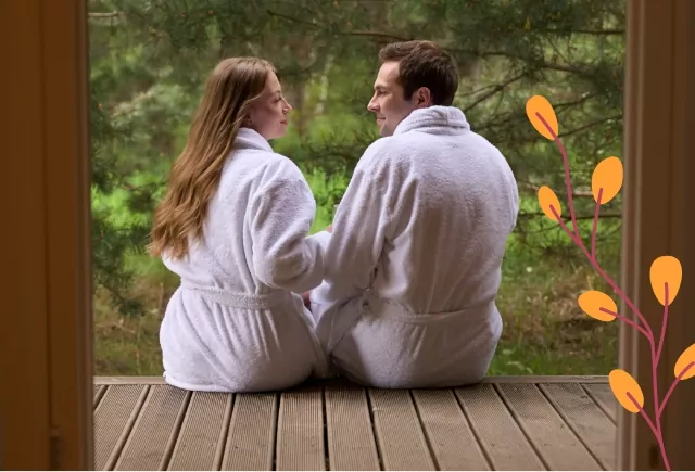 A couple in white bathrobes sitting on a wooden deck, gazing at each other with a peaceful forest setting in the background.