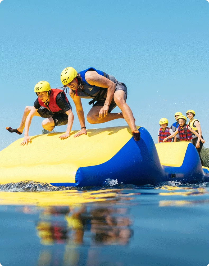 Two participants wearing helmets and life jackets climb over a large inflatable obstacle on a bright yellow and blue water course. In the background, a group of people waits for their turn, all wearing safety gear.