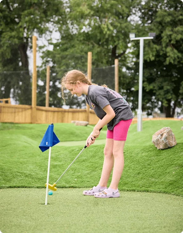 A young girl concentrating as she prepares to putt a golf ball on a mini-golf course. She is wearing a dark t-shirt, pink shorts, and Crocs with character charms, standing on artificial grass near a hole with a blue flag.
