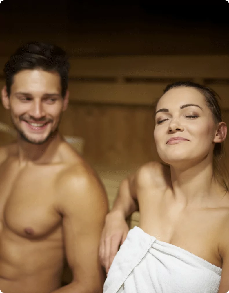 A man and a woman enjoying a relaxing sauna session. The woman, wrapped in a towel, leans back with her eyes closed, while the man smiles at her in the warm, wooden sauna setting.