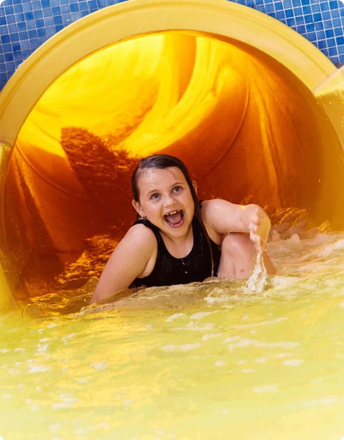 A young girl with wet hair and a big smile splashes out of a bright yellow water slide, wearing a black swimsuit. The water rushes around her as she enjoys the excitement of the splash zone.