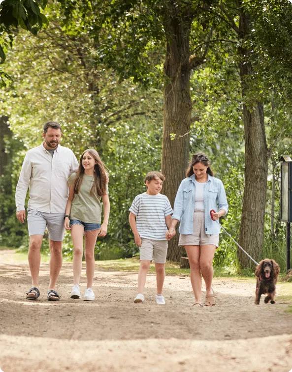 A family of four, along with their small brown dog, enjoying a walk through a lush green woodland trail. The parents and children are smiling and chatting as they stroll under the trees. The text ‘Woodland Walks’ is displayed at the bottom.
