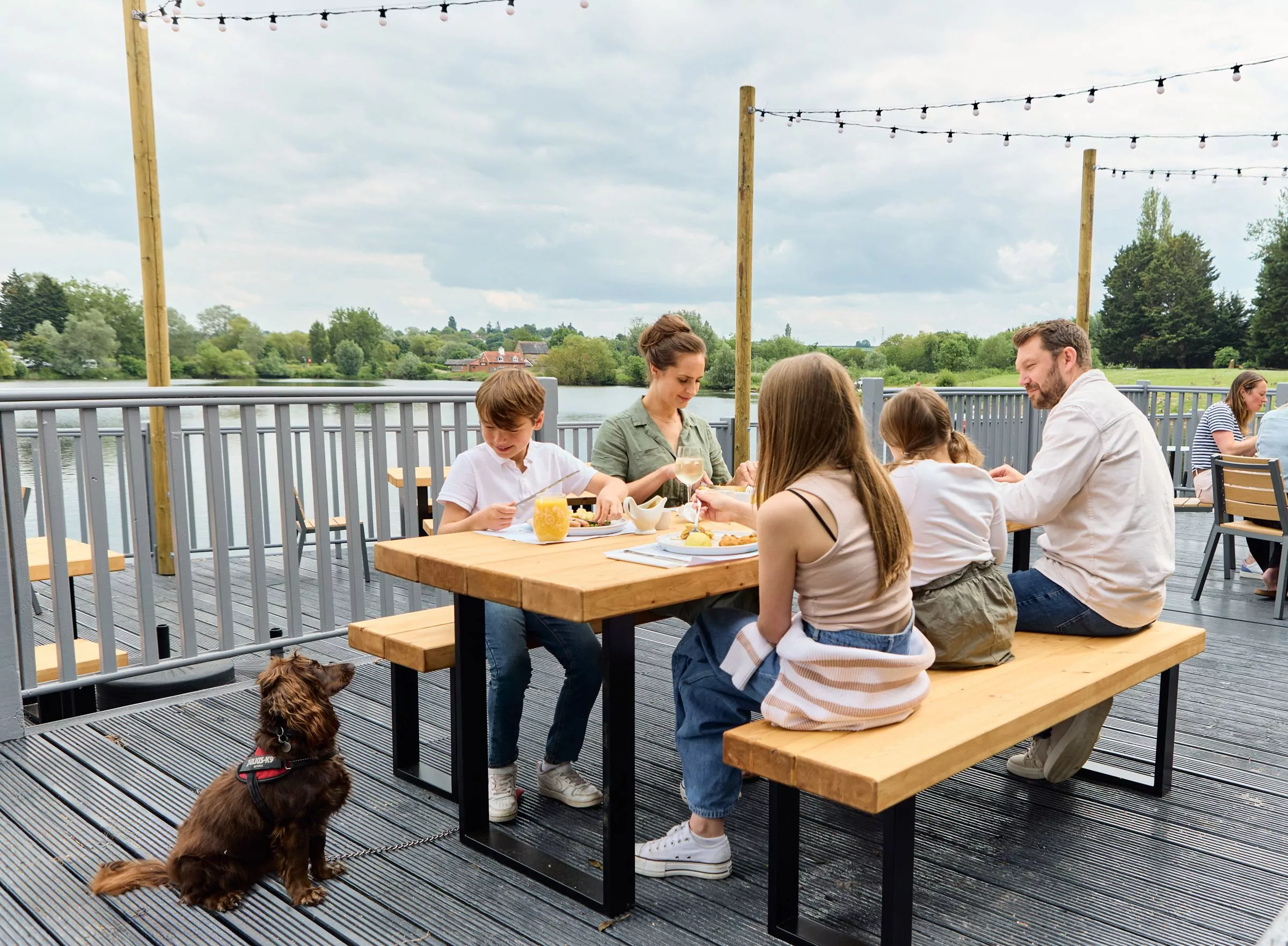 Family having a relaxed outdoor meal on a lakeside terrace with string lights above and a dog sitting nearby.