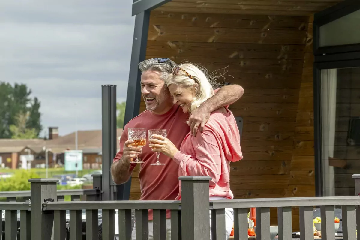 Smiling couple enjoying drinks together on the decking of a modern wooden lodge at Meadow Bay Villages
