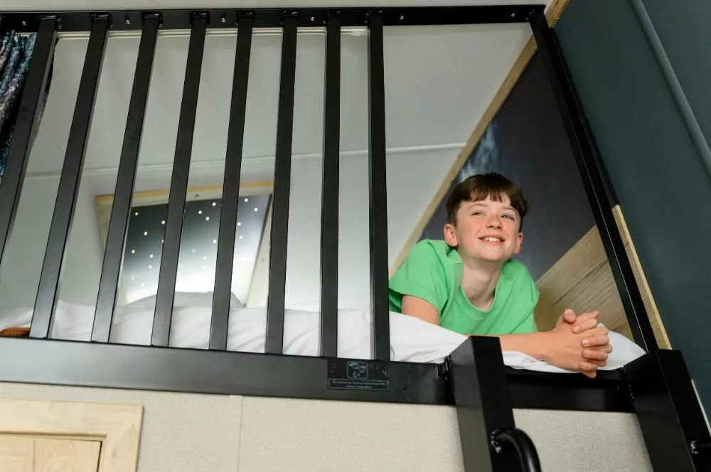 Smiling child lying in a raised loft-style bed with safety bars inside a modern holiday caravan at Meadow Bay Villages