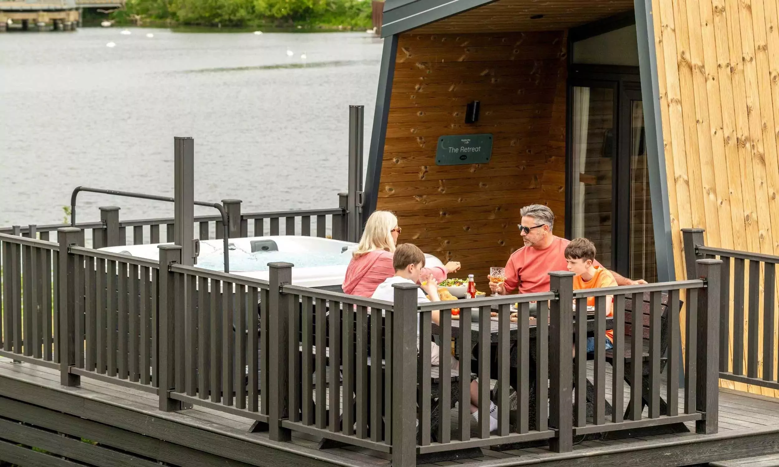 Family enjoying outdoor dining beside a hot tub on the decking of a lakeside lodge at Billing Aquadrome, part of Meadow Bay Villages.