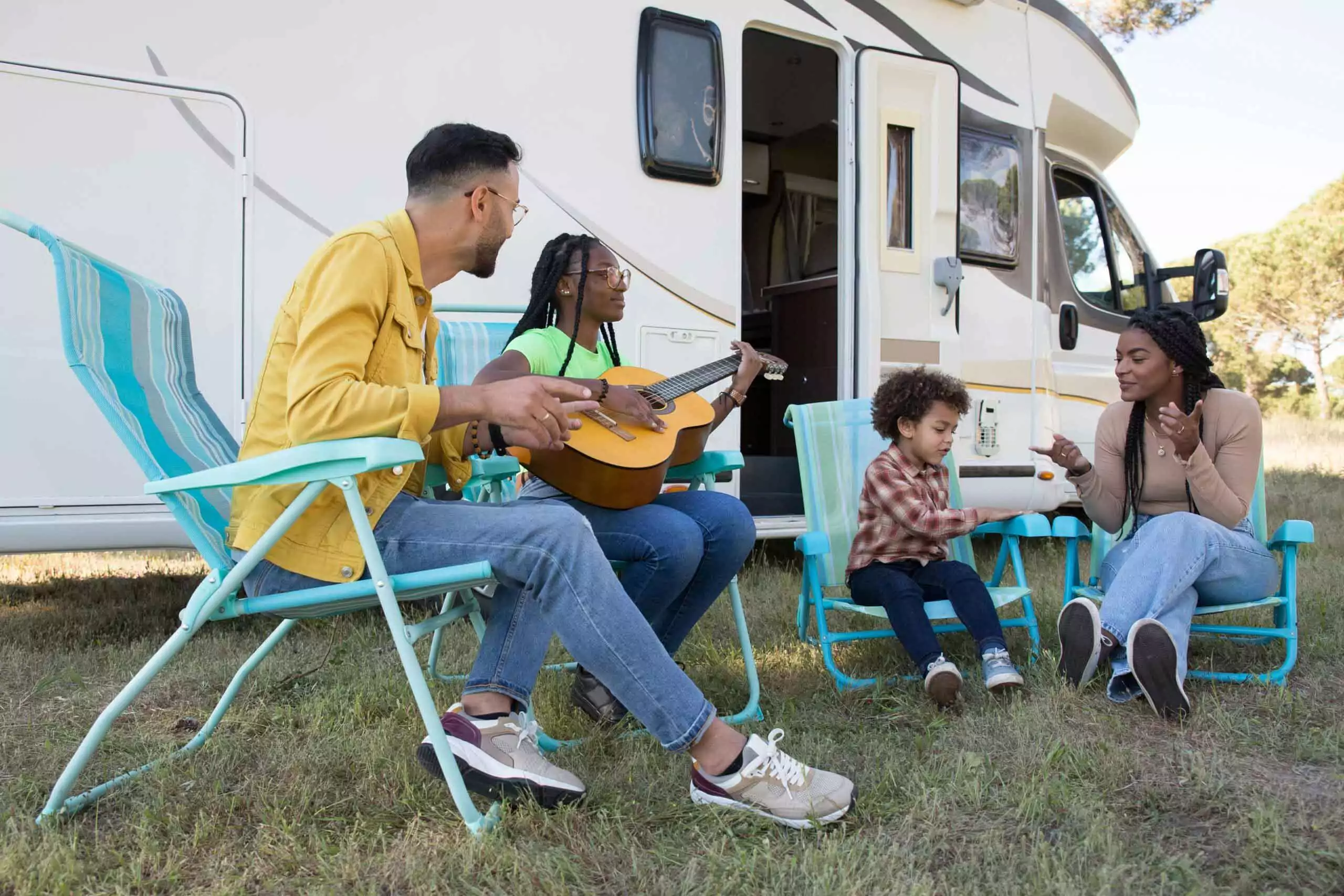 Family relaxing outside their caravan, playing guitar and chatting on fold-out chairs at Meadow Bay Villages
