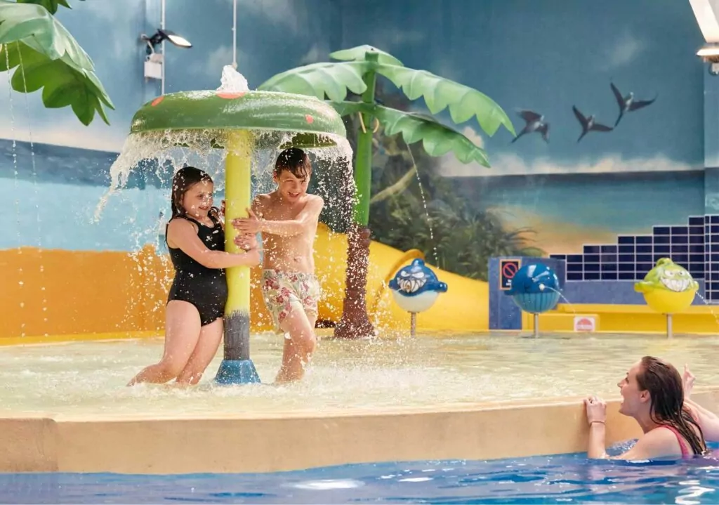 Children playing under a water feature in the indoor splash zone at Meadow Bay Villages’ Billing Aquadrome, with tropical-themed decor and shallow pool