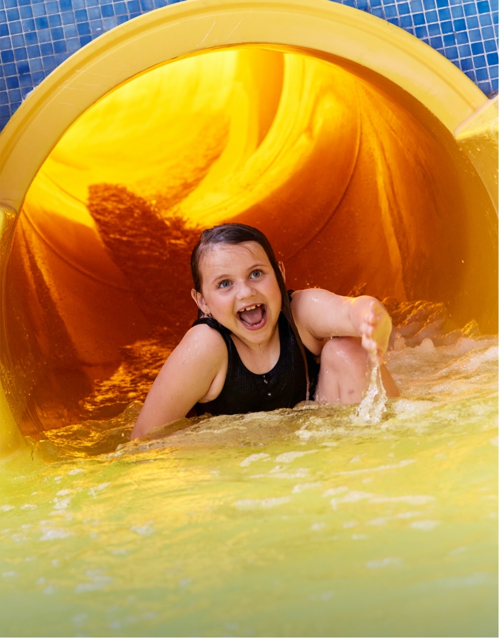 Smiling child splashing into the pool after riding down a yellow water slide at Billing Aquadrome