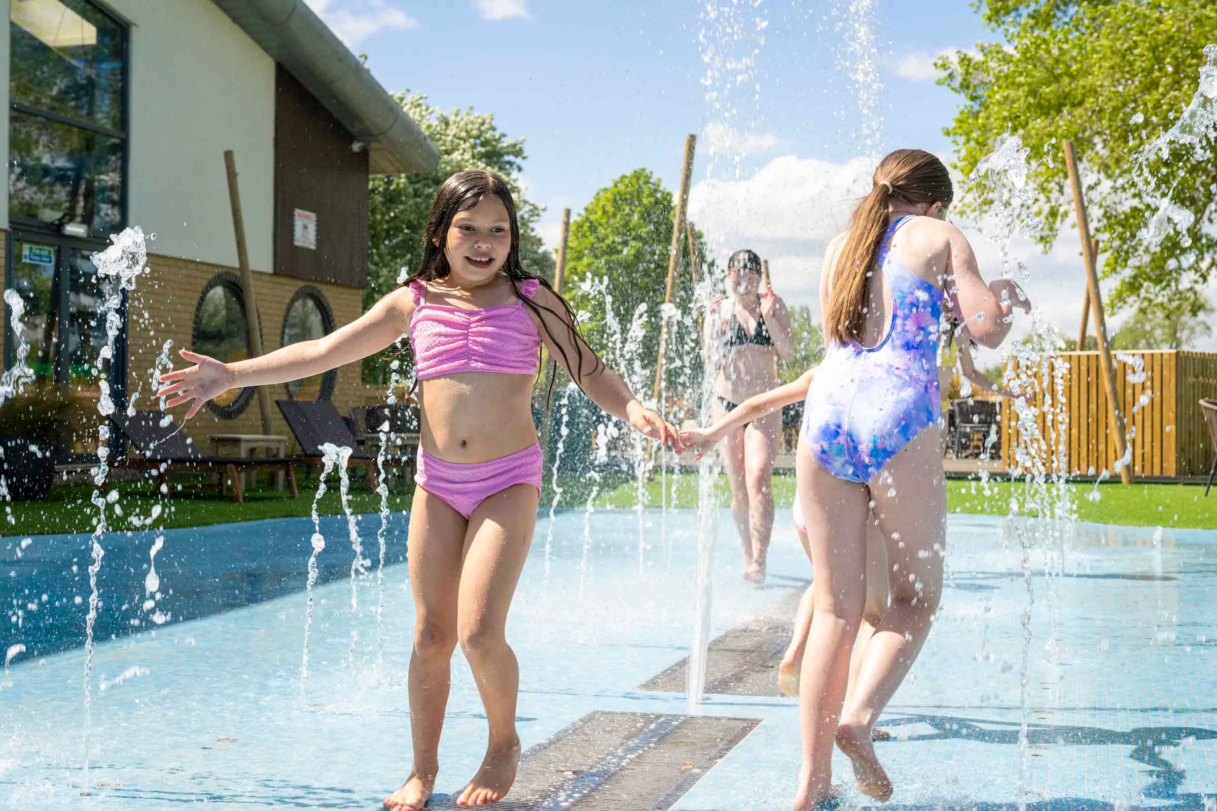 Children playing and laughing in an outdoor splash pad area with water jets under a sunny sky at Meadow Bay Villages.