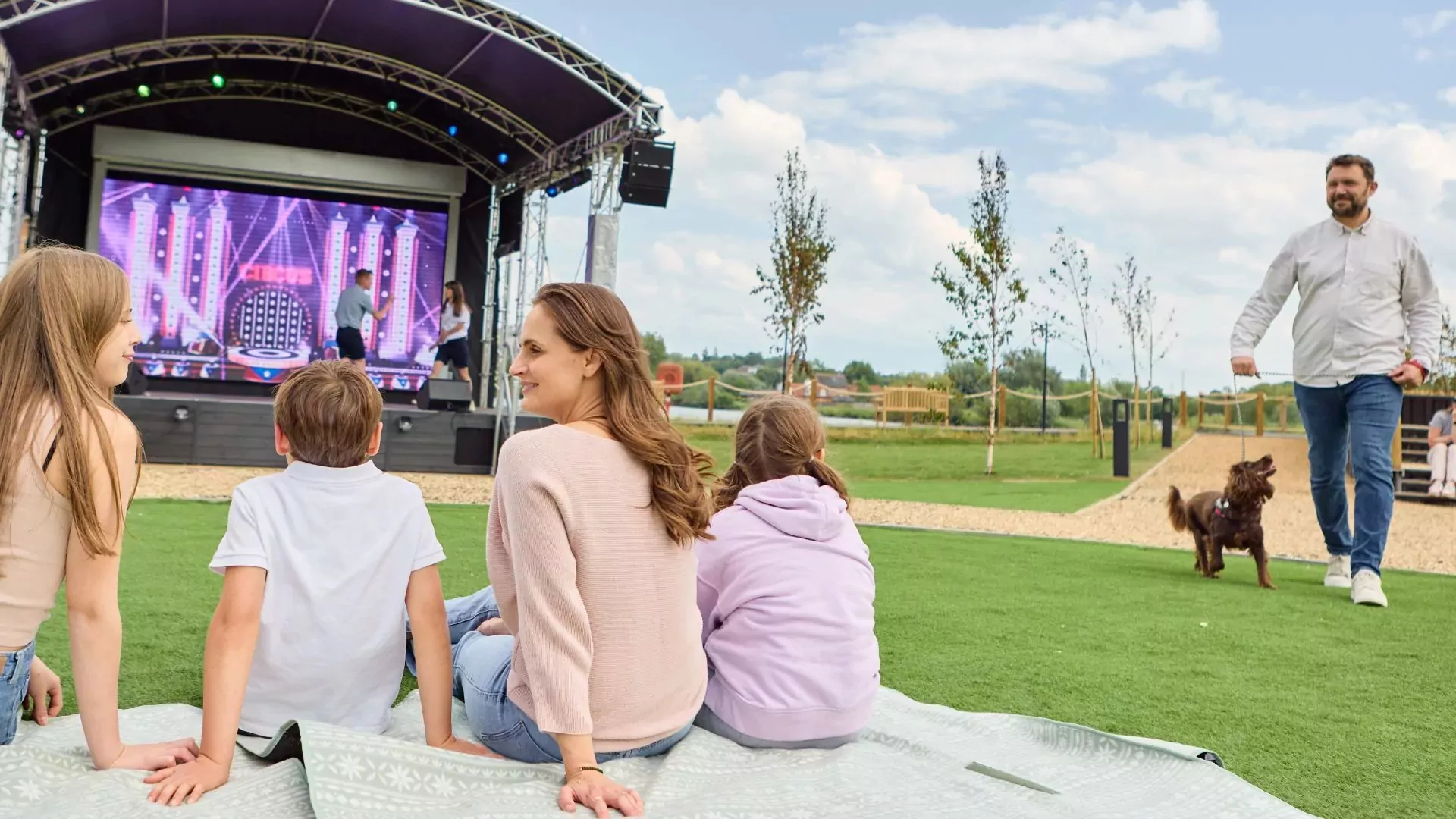 Family enjoying outdoor live entertainment at Meadow Bay Villages, with a dog walker passing by in the background