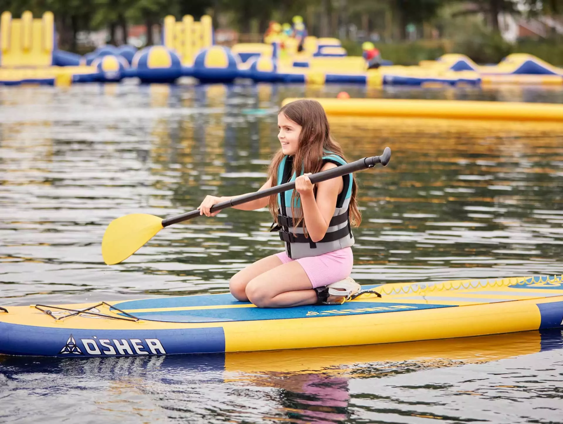 Young girl kneeling on a paddleboard with a yellow paddle at Meadow Bay Villages, with inflatable aqua park in the background