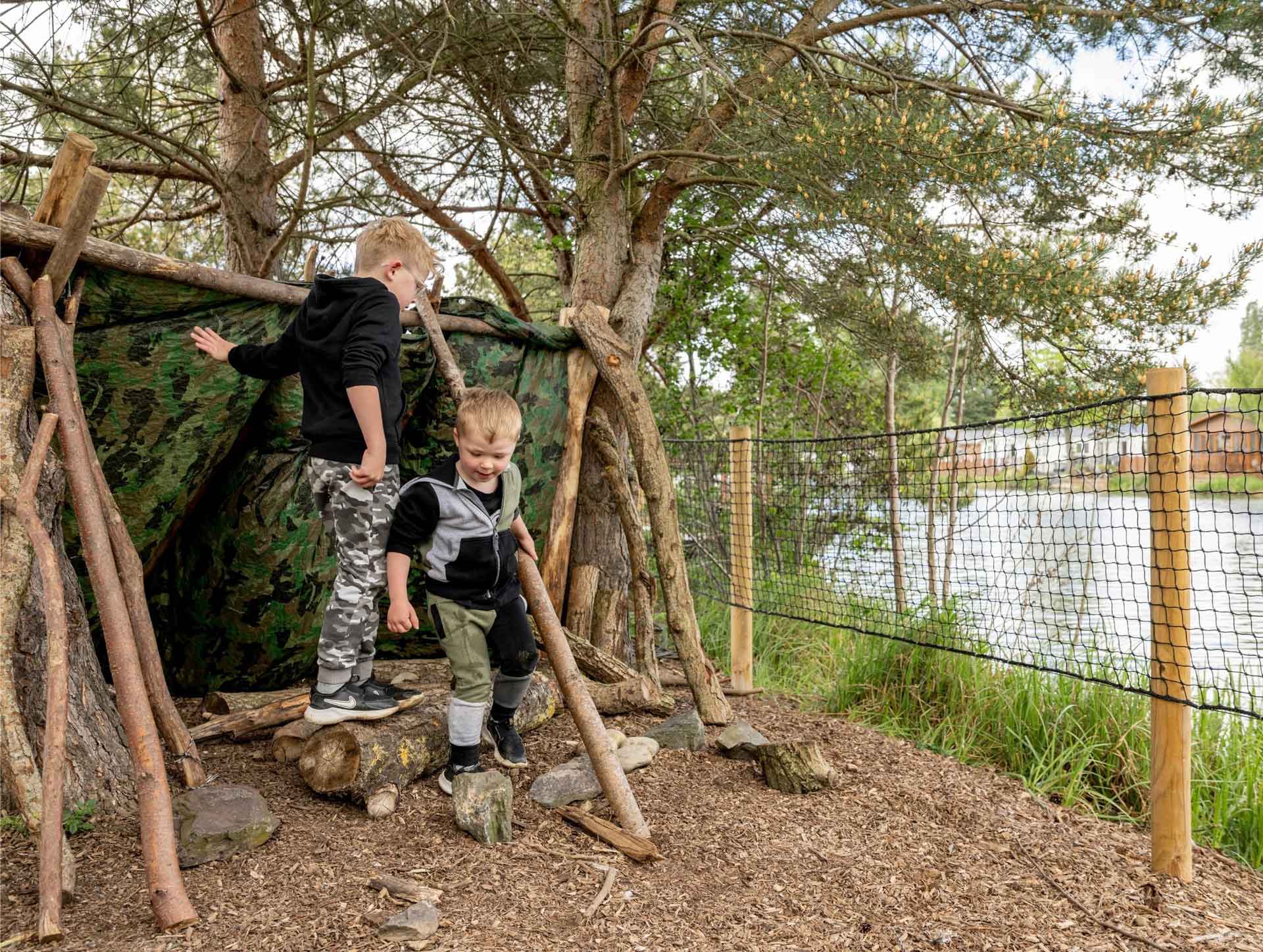 Two young children playing in a handmade woodland den made from logs and camo tarp at Meadow Bay Villages