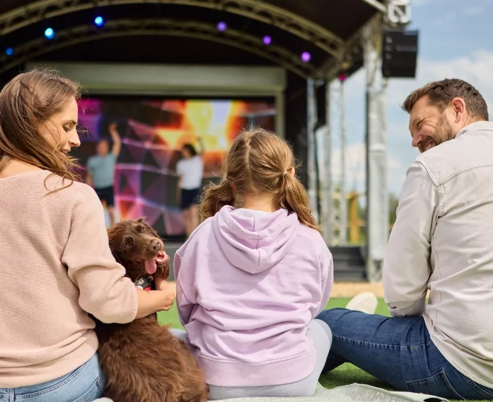 Family sitting on the grass with their dog enjoying a live outdoor show at Meadow Bay Villages’ Billing Aquadrome, with colourful stage lights in the background