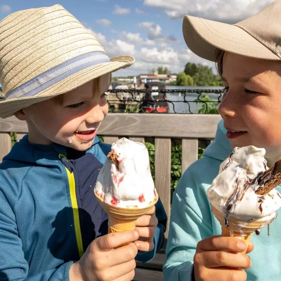 Two young boys in sun hats enjoying melting ice cream cones while sitting on a bench at Meadow Bay Villages’ Billing Aquadrome on a sunny day