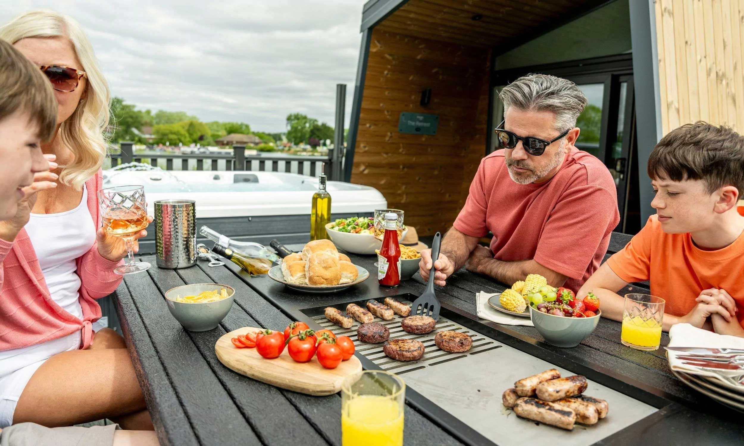 Family enjoying a summer BBQ with drinks beside a hot tub on the decking of their holiday lodge at Billing Aquadrome, part of Meadow Bay Villages.
