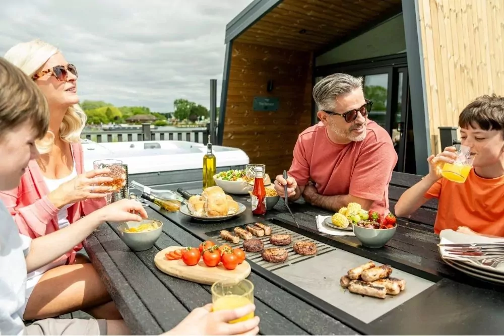 Family enjoying a barbecue lunch together on the terrace of a modern holiday lodge at Meadow Bay Villages’ Billing Aquadrome, with grilled food and summer drinks