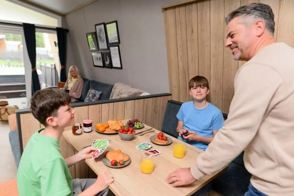 Family enjoying quality time and snacks around the dining table in a modern holiday home at Meadow Bay Villages’ Billing Aquadrome, with card games and fresh fruit