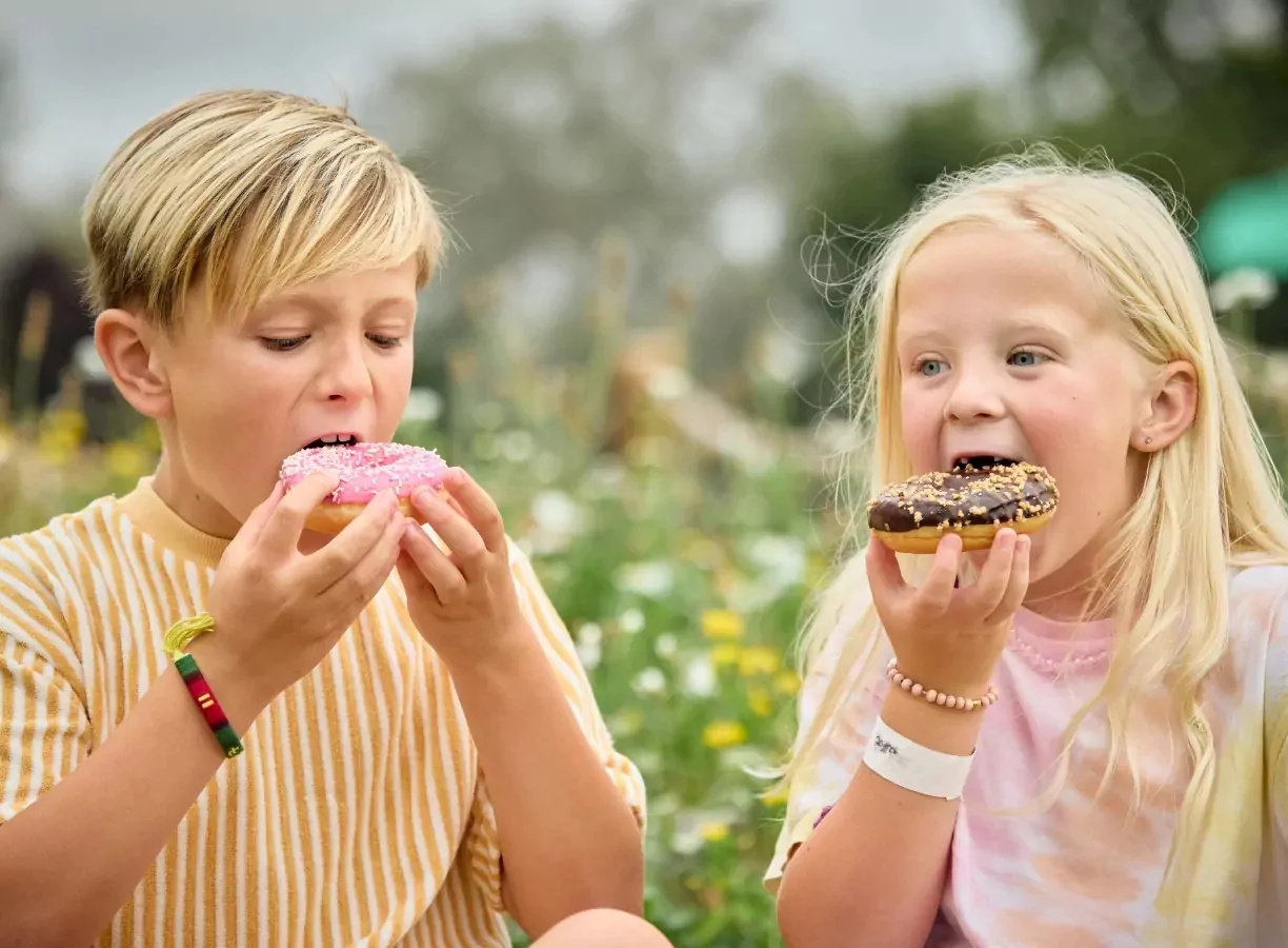 Two children enjoying colourful doughnuts outdoors at Meadow Bay Villages, surrounded by wildflowers