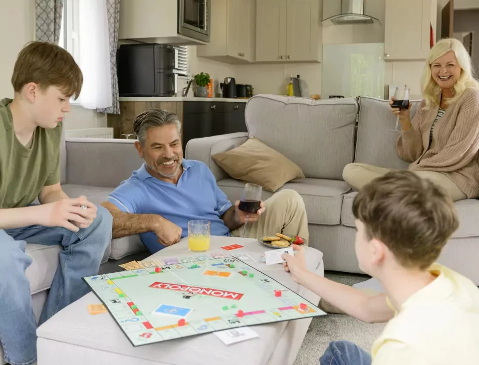 Family enjoying a game of Monopoly together in a modern caravan lounge at Meadow Bay Villages, sharing drinks and snacks