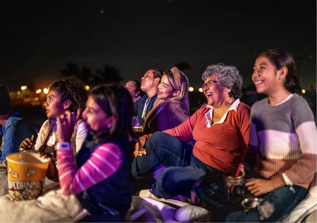 A multigenerational family enjoying an outdoor cinema night at Meadow Bay Villages. Everyone is smiling and laughing together while watching a film under the stars. A bucket of popcorn and drinks are visible, adding to the cozy, fun-filled evening atmosphere.