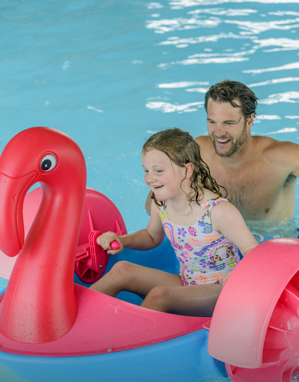 A young girl enjoying a ride on a pink flamingo paddle boat in the swimming pool with her dad at Billing Aquadrome.