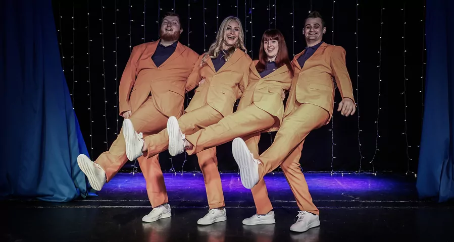 Four performers in coordinated orange suits dancing on stage during the Tribe Takeover show at Billing Aquadrome
