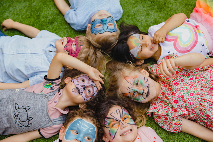 Group of children lying on the grass with colourful face paint designs during a fun outdoor activity at Billing Aquadrome