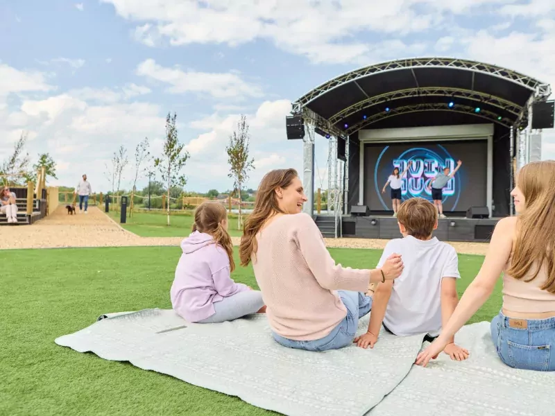 Family sitting on a picnic blanket watching a live outdoor stage performance during an entertainment event at Billing Aquadrome