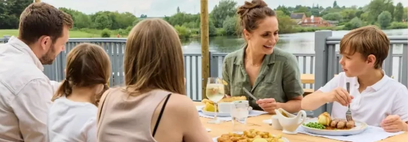A group enjoys a meal outdoors by a river, featuring plates of food and drinks, with green trees in the background.