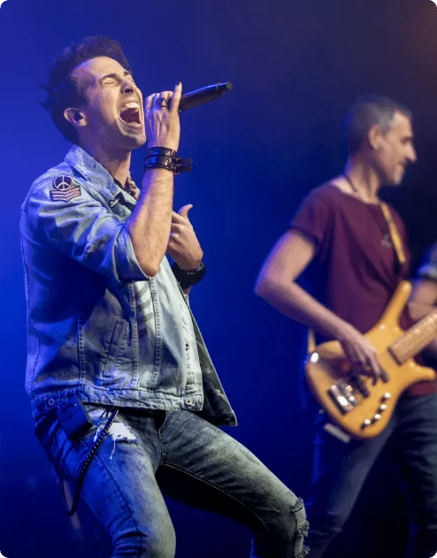 A man singing into a microphone whilst his band mate plays the guitar behind him