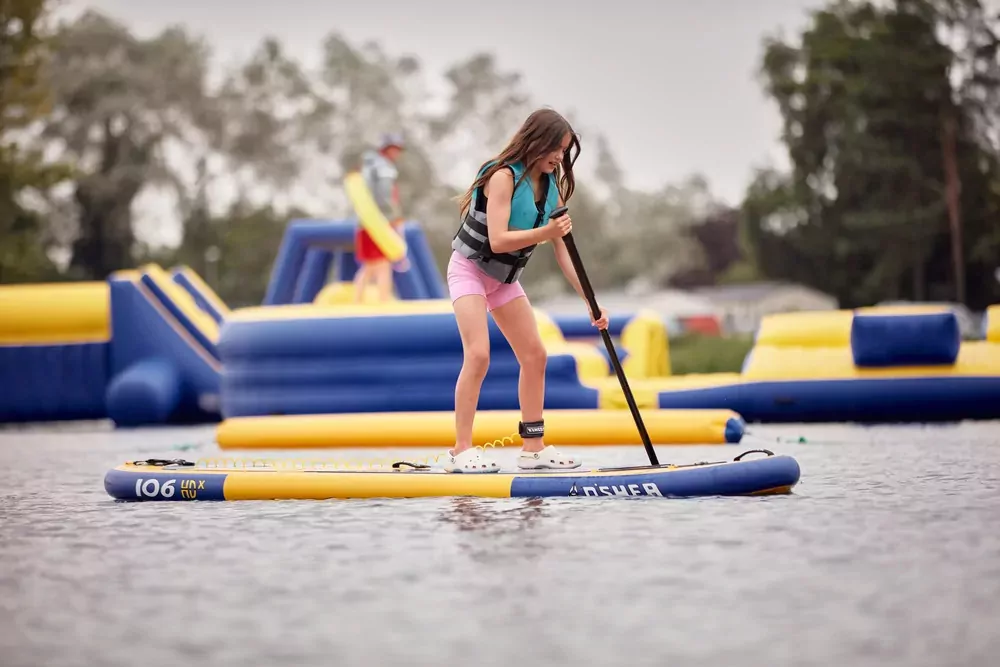 Girl wearing a life jacket paddleboarding on the lake with inflatable obstacles in the background at Billing Aquadrome