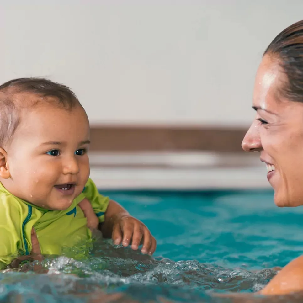 Smiling baby in a neon green swimsuit enjoying a swim with a parent in a pool.