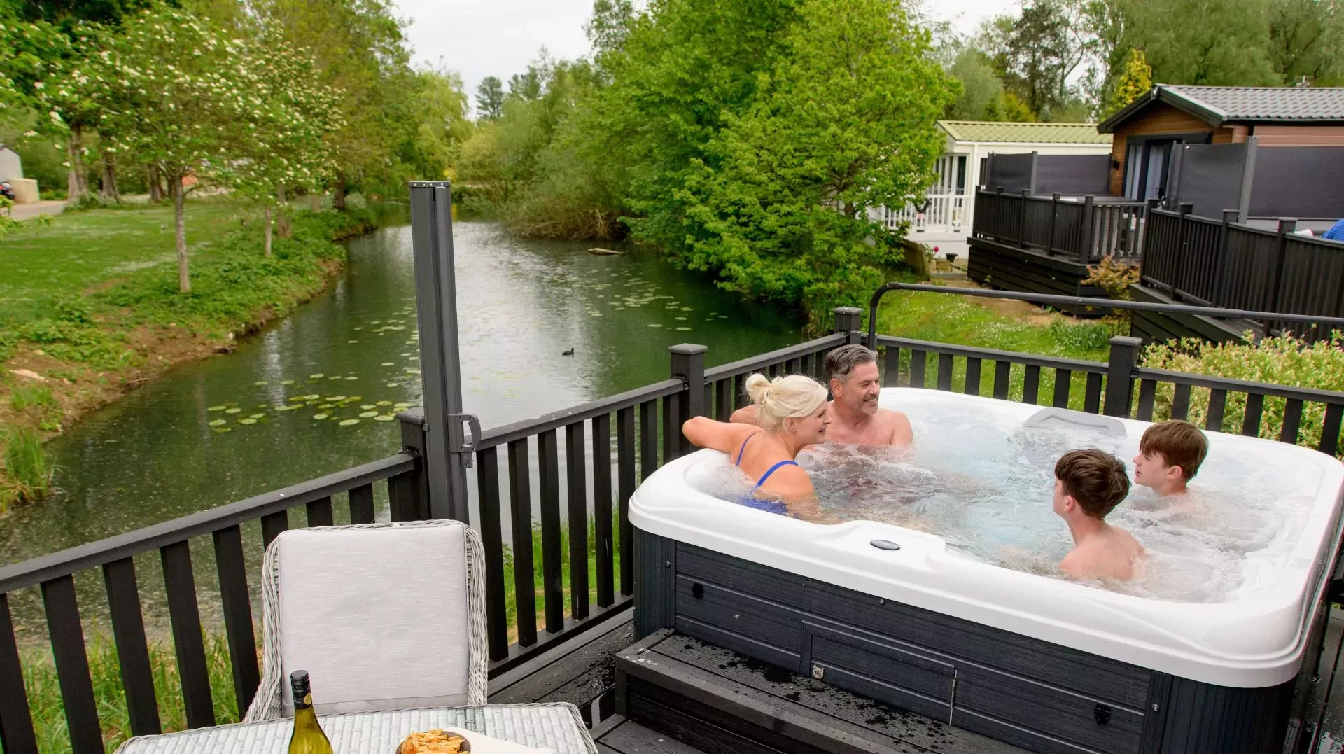 Scenic view of a family enjoying a hot tub on the decking of a lakeside lodge at Billing Aquadrome holiday park, with drinks and snacks nearby