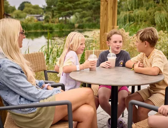 Children enjoying milkshakes and chatting at a lakeside café table at Meadow Bay Villages holiday park, surrounded by nature.