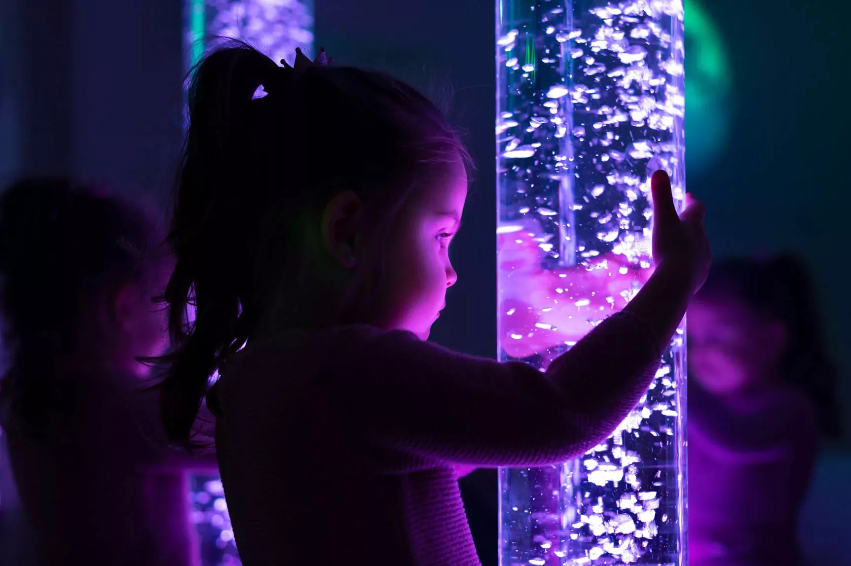 Young child interacting with a glowing purple sensory bubble tube in the darkened Zen Den at Meadow Bay Villages