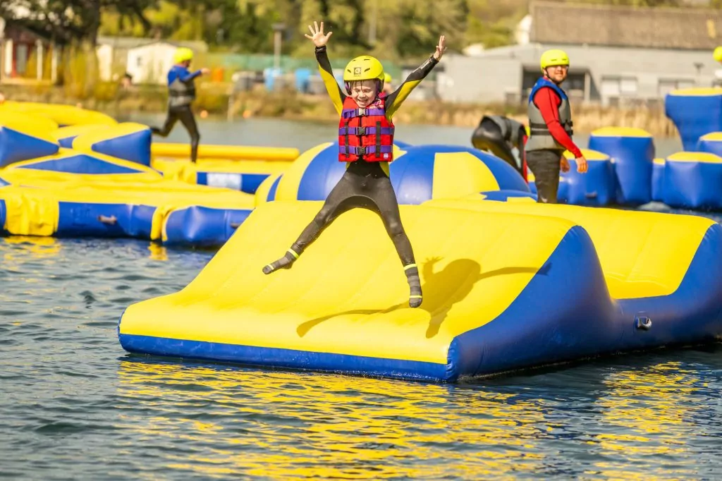 A young boy in a wetsuit, life jacket, and safety helmet jumping with excitement on an outdoor inflatable aqua park course at Billing Aquadrome