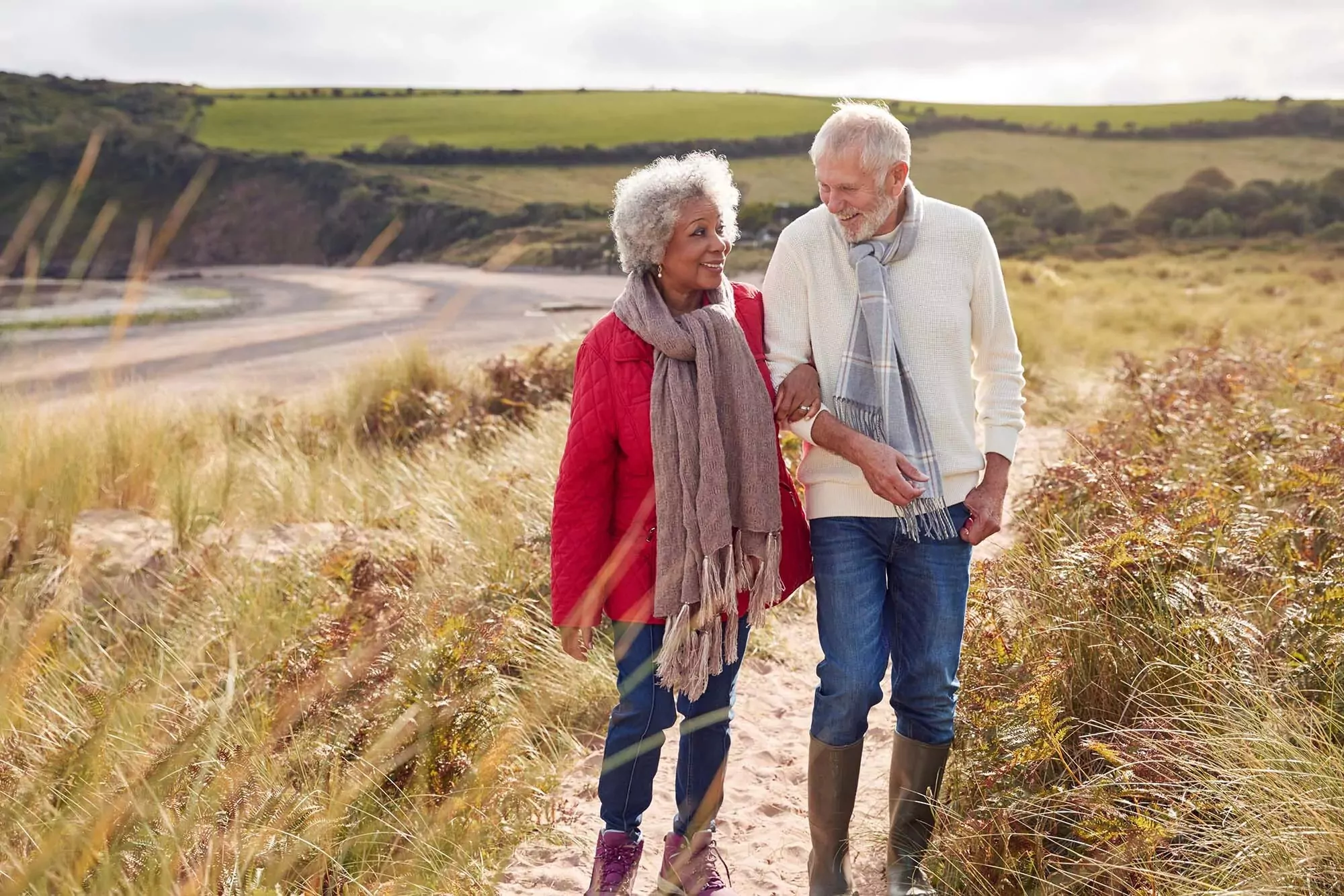 Smiling couple enjoying an autumn walk along a coastal path near Meadow Bay Villages' Golden Leas, wrapped in scarves and surrounded by golden grasses.