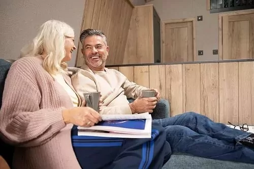 Smiling couple relaxing on a sofa with hot drinks inside a modern lodge at Golden Leas.