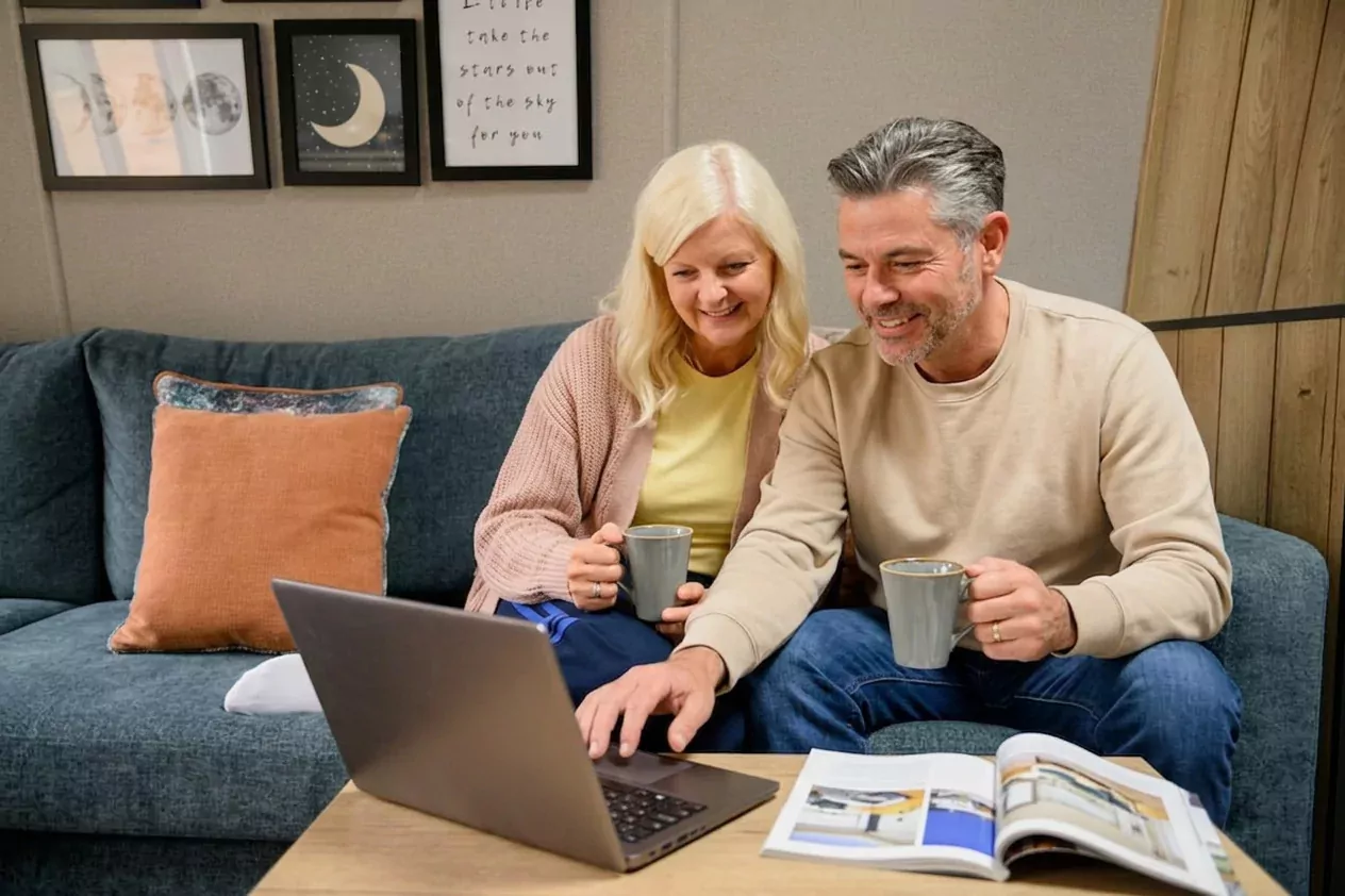 A smiling senior couple sitting on a blue sofa, holding mugs, and planning on a laptop with an open magazine at Golden Leas