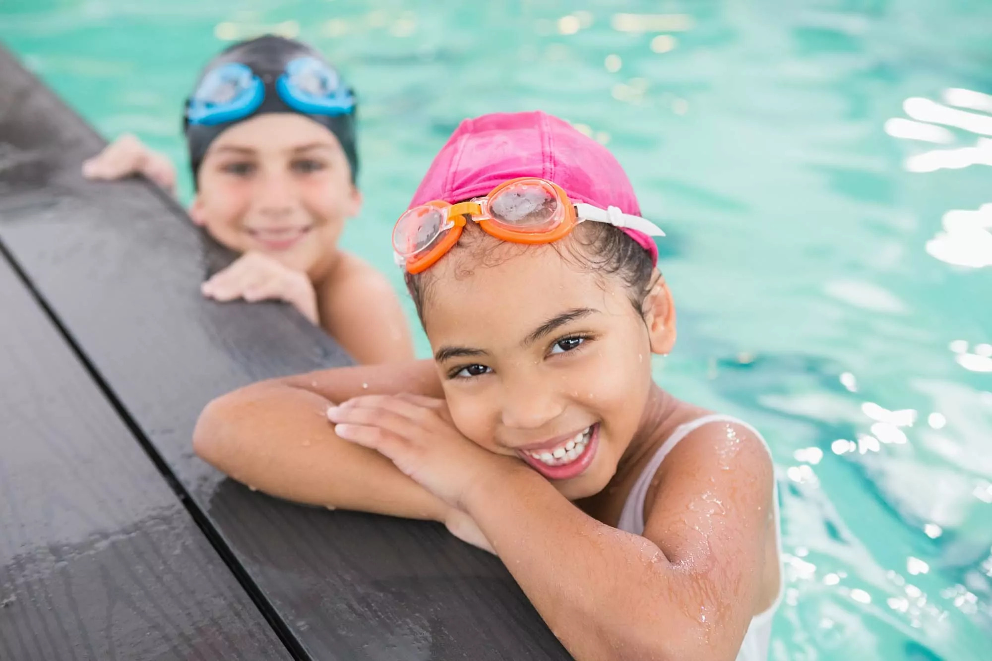 Two smiling children enjoying a swim on Hayling Island, with one girl in a pink swim cap and goggles resting at the pool edge and a boy in a blue cap in the background.