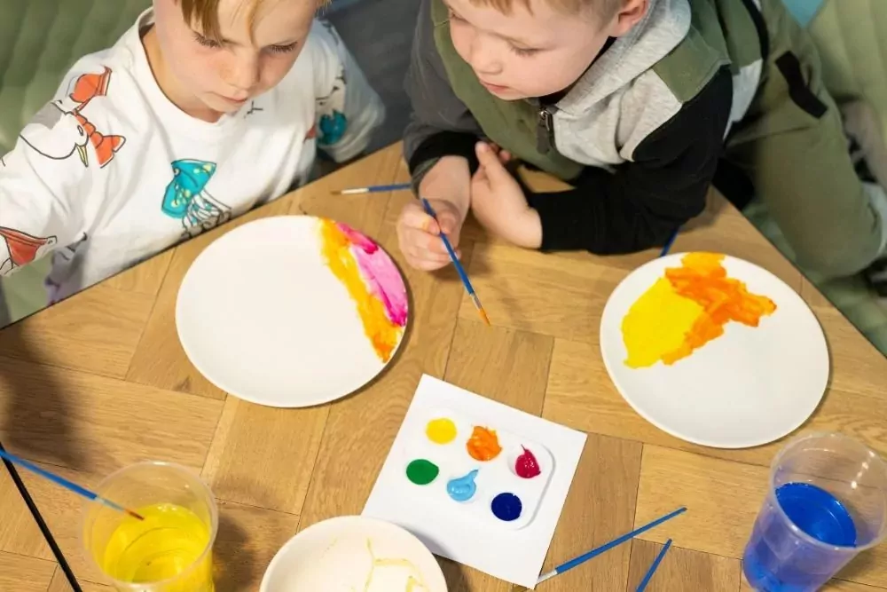 Two young boys painting white plates with bright colours using paintbrushes and small pots of paint during a creative activity at Hayling Island.