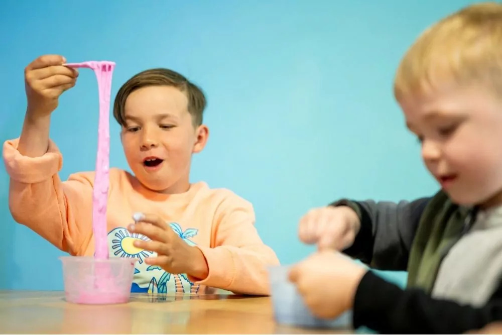 Two young boys enjoying a slime activity, with one stretching bright pink slime and the other focused on stirring his own container, against a light blue background at Hayling Island.
