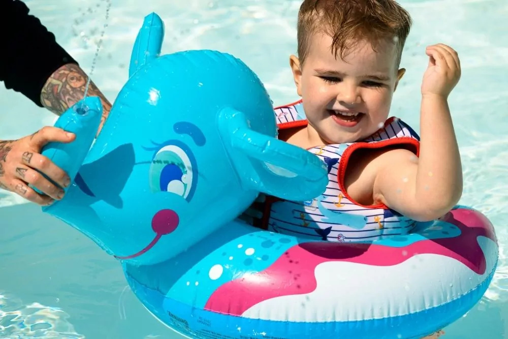 Happy toddler floating in a colourful inflatable ring shaped like a blue animal, splashing and smiling in the shallow pool on Hayling Island.