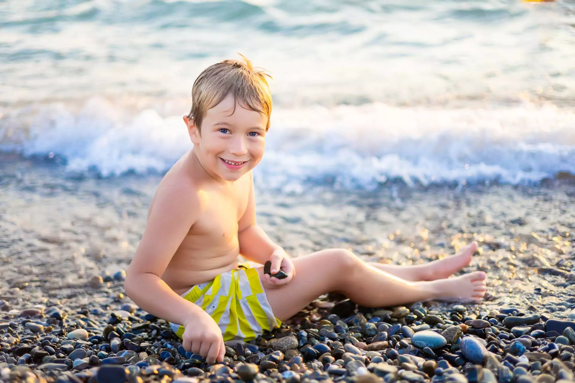A happy child sitting on a pebble beach, wearing yellow striped swim shorts, smiling at the camera with ocean waves gently rolling onto the shore in the background.