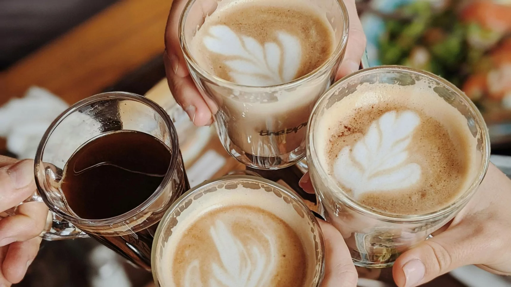 A group of friends toasting with glass cups of coffee, featuring beautifully crafted latte art and a black coffee, during a cozy brunch setting.
