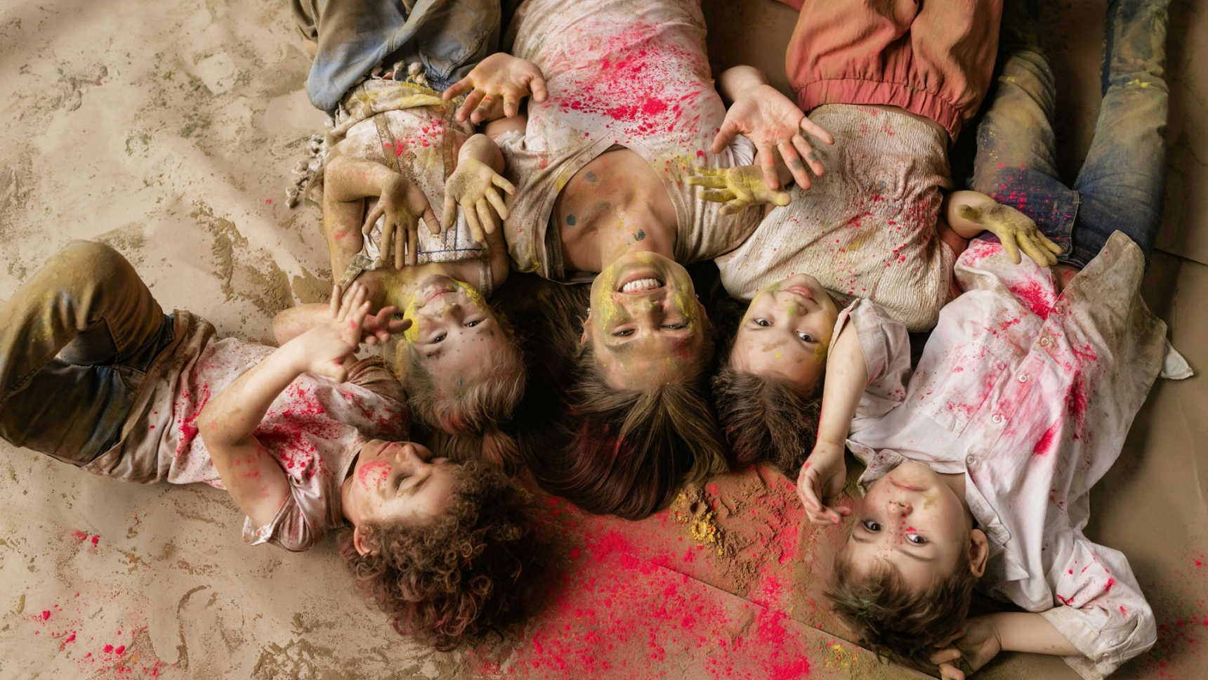 A joyful group of children and an adult lying on the ground covered in colorful powder, laughing and playing during a vibrant color throw party.